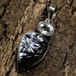 Handcrafted silver pendant featuring a dark metallic silicon stone with natural reflective patterns and a clear rock crystal accent in a polished silver setting on textured wood background.
