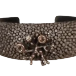 Handcrafted stingray leather bracelet with textured dark surface, silver coral-inspired centerpiece, and green peridot gemstones displayed on a crystal stand.