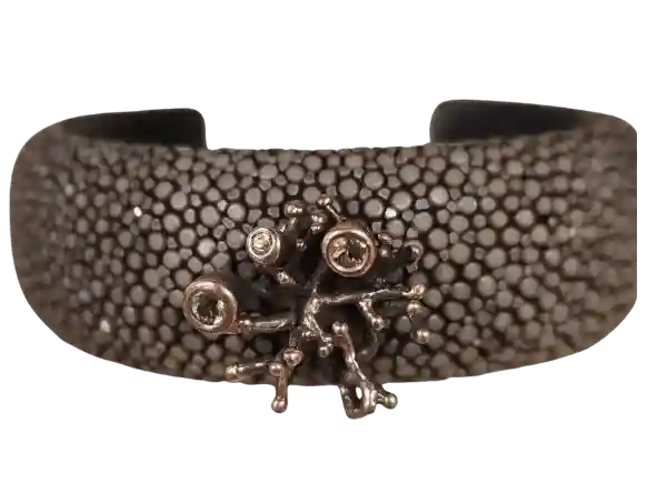 Handcrafted stingray leather bracelet with textured dark surface, silver coral-inspired centerpiece, and green peridot gemstones displayed on a crystal stand.