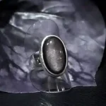 Handcrafted sterling silver ring featuring a natural oval Silver Sheen Obsidian gemstone from the Canary Islands displayed on a clear acrylic stand.