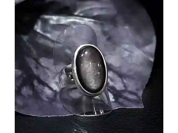 Handcrafted sterling silver ring featuring a natural oval Silver Sheen Obsidian gemstone from the Canary Islands displayed on a clear acrylic stand.